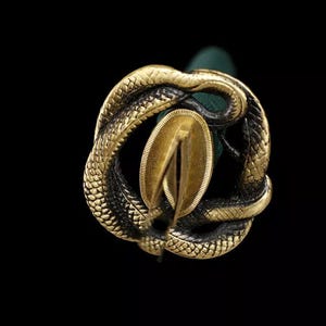 May include: Close-up of a decorative sword hilt with a gold-colored snake design. The snake is coiled around a gold-colored oval-shaped element. The background is black.