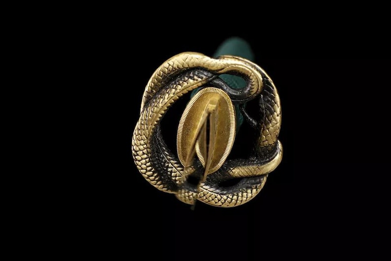 May include: Close-up of a decorative sword hilt with a gold-colored snake design. The snake is coiled around a gold-colored oval-shaped element. The background is black.