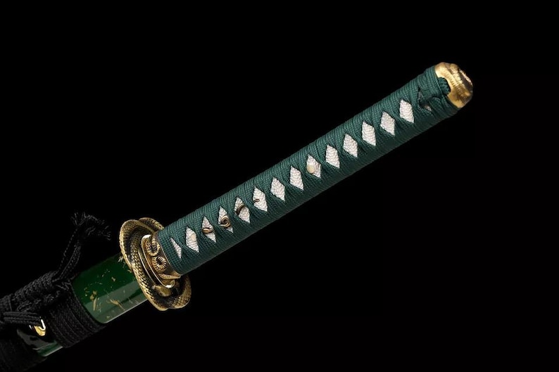 May include: Close-up of a katana handle with a dark green wrap and white diamond-shaped accents. The hilt features gold-colored metal detailing and a black cord. The end cap is gold. The background is black.
