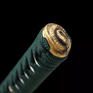 May include: Close-up of a katana handle. The handle is wrapped in dark green fabric and features a gold-colored metal pommel with a detailed snake design. The background is black.