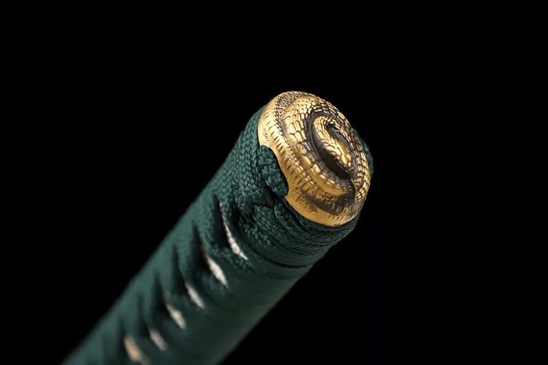 May include: Close-up of a katana handle. The handle is wrapped in dark green fabric and features a gold-colored metal pommel with a detailed snake design. The background is black.