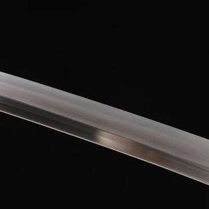 May include: Close-up of a polished, silver-colored sword blade against a black background. The blade is angled diagonally, reflecting light and showcasing its sharp edge. The image highlights the blade's metallic sheen.