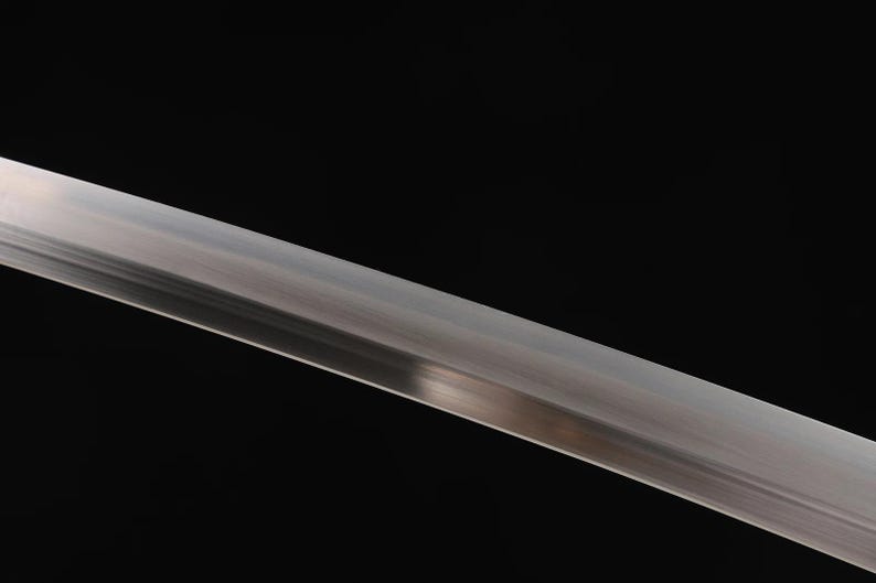 May include: Close-up of a polished, silver-colored sword blade against a black background. The blade is angled diagonally, reflecting light and showcasing its sharp edge. The image highlights the blade's metallic sheen.