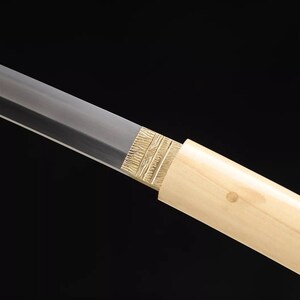 May include: Close-up of a Japanese katana sword. The blade is silver, and the handle is wrapped in a light-colored wood. The guard is gold-colored with a textured design. The background is black.