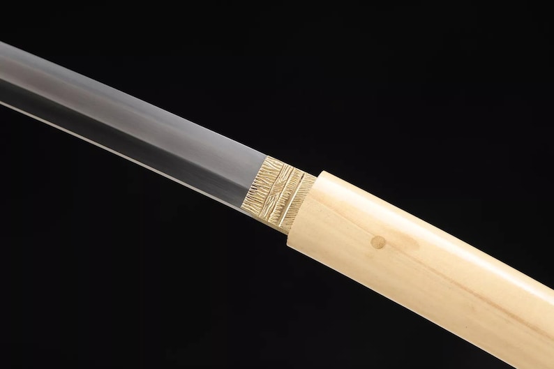 May include: Close-up of a Japanese katana sword. The blade is silver, and the handle is wrapped in a light-colored wood. The guard is gold-colored with a textured design. The background is black.