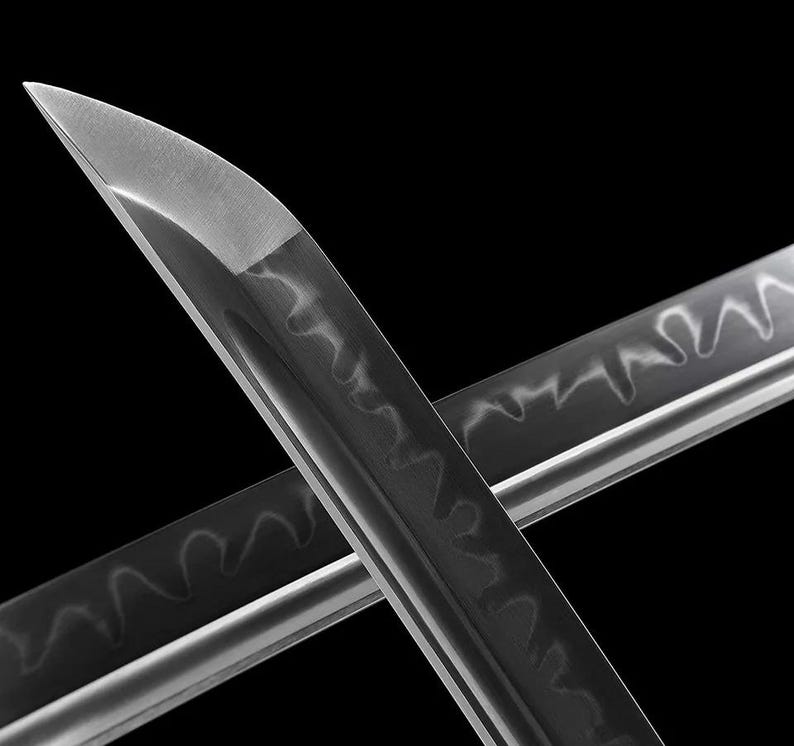 May include: Close-up of two crossed katana blades. The blades are silver with a dark, wavy pattern along the edge. The tips are sharp and angled, reflecting light against a black background.