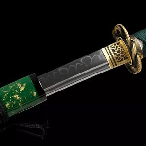 May include: Close-up of a katana sword with a black blade and a green handle. The handle is wrapped in black cord and features gold accents. The blade has a unique pattern, and the guard is gold.