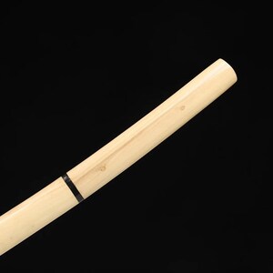 May include: Angled view of a light-colored wooden sword handle against a black background. The handle has a natural wood grain and a black band near the center. The end of the handle is rounded.