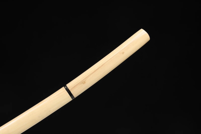 May include: Angled view of a light-colored wooden sword handle against a black background. The handle has a natural wood grain and a black band near the center. The end of the handle is rounded.