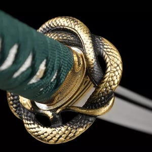 May include: Close-up of a katana sword hilt. The hilt features a gold-colored snake design and a green handle wrap. The blade is visible in the background. The design is set against a black background.