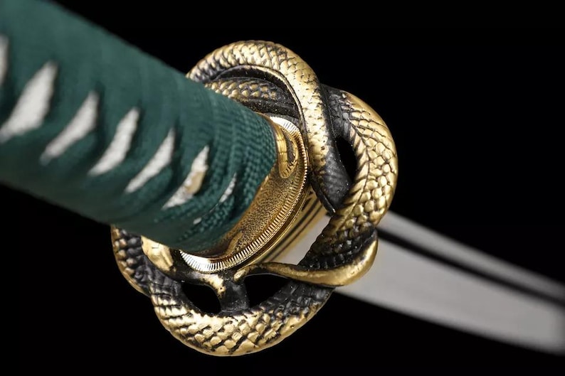 May include: Close-up of a katana sword hilt. The hilt features a gold-colored snake design and a green handle wrap. The blade is visible in the background. The design is set against a black background.