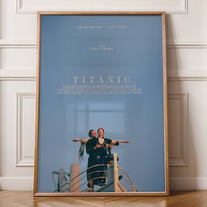 Op de afbeelding: Ingelijste filmposter voor de film "Titanic" met Leonardo DiCaprio en Kate Winslet. De poster heeft een blauwe achtergrond met de titel en credits in gouden letters. De afbeelding toont de iconische scène van de twee personages met hun armen uitgestrekt.