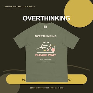 May include: An olive green t-shirt featuring a graphic of a snail with a loading icon above it. The text reads OVERTHINKING, PLEASE WAIT, and STILL PROCESSING. The design includes a dashed circular line, curved lines, and a pink snail face.