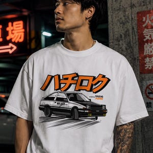 Könnte beinhalten: Ein weißes Kurzarm-T-Shirt mit einer grafischen Illustration eines weiß-schwarzen Toyota AE86 Trueno Autos. Über dem Auto ist der japanische Text "ハチロク" in fetten orangefarbenen Buchstaben mit schwarzer Umrandung gedruckt. Rechts neben dem Auto steht der Text "TRUENO AE86" in kleinerer schwarzer Schrift.