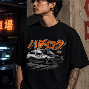 May include: A black short-sleeved t-shirt featuring a graphic print of a white and black AE86 car. Above the car, the Japanese text "ハチロク" is printed in bold orange letters with a black outline. The design includes the text "TRUENO AE86" in small orange letters.