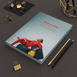 May include: A hardcover notebook with the word "hammertime" and its definition printed on the cover. The cover features a photo of a person in a red racing suit. Gold and black pencils and binder clips are also visible.