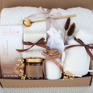 May include: A brown gift box filled with a white blanket, a white mug, a bar of soap, a jar of candle, a small bottle of lotion, a bag of bath salts, and a chocolate bar that says "Le Belge Napa Valley Hazelnut Coffee Milk Chocolate Net Wt 3 oz (85g)".