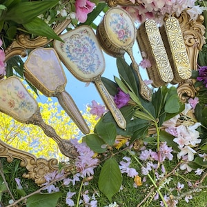 Könnte beinhalten: Ein verzierter goldgerahmter Spiegel zeigt eine Sammlung von Vintage-Kosmetikartikeln. Das Set umfasst Handspiegel und Bürsten mit floralen Designs und goldenen Akzenten. Die Anordnung ist von rosa und lila Blumen und Grün umgeben, was eine romantische Ästhetik schafft.