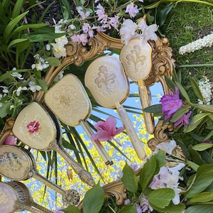 Könnte beinhalten: Ein antikes Schminkset mit einem goldgerahmten Spiegel und mehreren passenden Handspiegeln und Bürsten. Das Set ist von Grün und Blumen umgeben und schafft eine Vintage-Ästhetik. Die Handspiegel weisen verzierte Designs und florale Akzente auf.