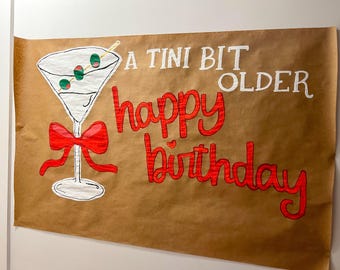 May include: A brown paper banner with a hand-drawn martini glass, red bow, and the words "A Tini Bit Older Happy Birthday" in white and red. The martini has green olives.