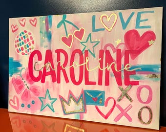 May include: A pink and white canvas with the name "CAROLINE" written in large pink letters. The canvas is decorated with hearts, stars, a crown, a butterfly, and the word "LOVE" in gold letters.
