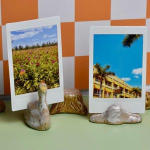 Op de afbeelding: Drie polaroid fotohouders met keramische voetjes. De houders tonen foto's van een gebouw, een bloemenveld en een gebouw met palmbomen. De voetjes hebben de vorm van een boot, een gans en een wolk.