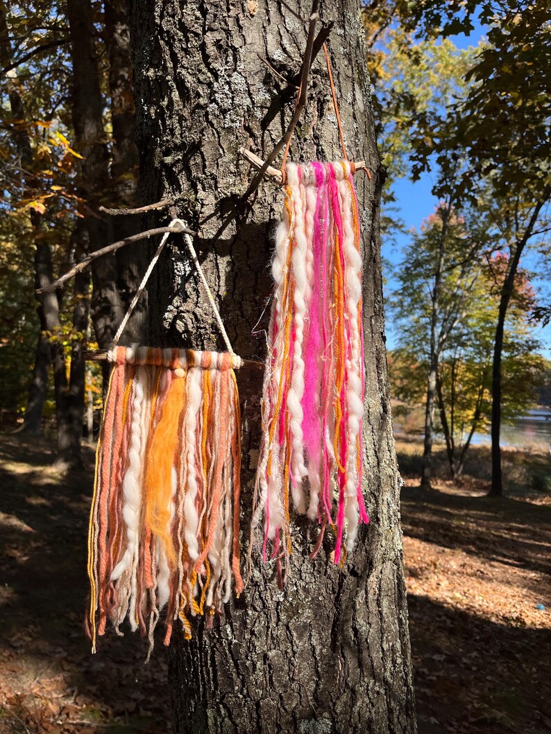 May include: Two handmade yarn wall hangings are displayed on a tree trunk. The larger hanging features vibrant pink, orange, and white yarn strands, while the smaller one showcases a palette of peach, cream, and gold yarn. Both hangings are woven with natural wood branches, creating a rustic, bohemian aesthetic. These unique textile art pieces would add a touch of warmth and color to any home decor.