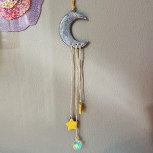 May include: A celestial-themed wall hanging featuring a crescent moon, yellow stars, and a crystal ball. The moon is a muted blue-gray, and the stars are a bright yellow. The crystal ball is clear with iridescent colors. The hanging is suspended by a natural-colored twine.