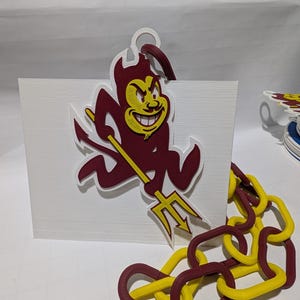 Puede incluir: Un logotipo de los Sun Devils de la Universidad Estatal de Arizona impreso en 3D en color granate y dorado con un tridente. El logotipo está unido a una base rectangular blanca. Una cadena de eslabones granate y dorados está en primer plano.