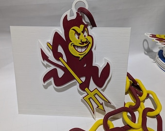 May include: A maroon and gold 3D-printed Arizona State University Sun Devils logo with a pitchfork. The logo is attached to a white rectangular base. A chain of maroon and gold links is in the foreground.