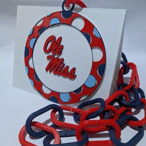 Puede incluir: Un letrero rectangular blanco con un diseño circular rojo, blanco y azul con las palabras "Ole Miss". Una cadena de eslabones rojos y azules está unida al letrero. El diseño tiene un patrón de lunares.