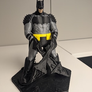 以下が含まれることがあります： 黒い六角形の台座の上に立つ、3Dプリントされたバットマンのアクションフィギュア。グレーのボディスーツ、黒いカウル、スパイク付きの黒い肩当て、黒いブーツ、鮮やかな黄色のユーティリティベルトを着用しています。フィギュアは黒いマントを前方に持っています。