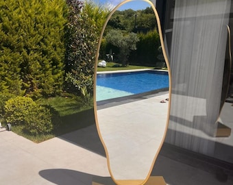 Op de afbeelding: Een goudkleurige, vrijvormige spiegel met een gouden basis. De spiegel reflecteert een zwembad en bomen.