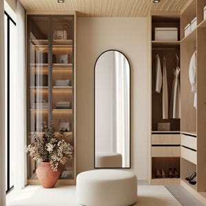 May include: A modern walk-in closet with light wood cabinetry, a full-length arched mirror, and a cream-colored ottoman. A large vase with dried flowers sits near a glass-fronted cabinet displaying handbags and folded clothing.