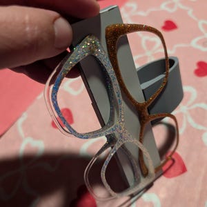K&ouml;nnte beinhalten: Zwei Brillenpaare mit Glitzer bedeckt. Ein Paar hat einen transparenten Rahmen mit blauem und silbernem Glitzer, das andere hat einen braunen Rahmen mit goldenem Glitzer. Die Brillen werden vor einem grauen Hintergrund gehalten.