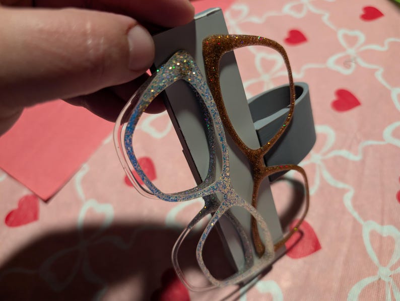 K&ouml;nnte beinhalten: Zwei Brillenpaare mit Glitzer bedeckt. Ein Paar hat einen transparenten Rahmen mit blauem und silbernem Glitzer, das andere hat einen braunen Rahmen mit goldenem Glitzer. Die Brillen werden vor einem grauen Hintergrund gehalten.