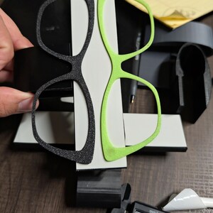 K&ouml;nnte beinhalten: Zwei Brillenrahmenpaare, eines in Schwarz und eines in Neongr&uuml;n, werden vor einem wei&szlig;en und schwarzen Hintergrund gezeigt. Der schwarze Rahmen hat ein strukturiertes, glitzerndes Aussehen. Ein digitaler Messschieber ist im Vordergrund zu sehen.