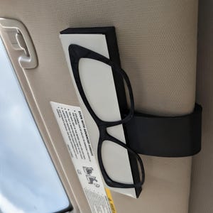 K&ouml;nnte beinhalten: Schwarze Brille mit Katzenaugen-Rahmen wird in einem schwarzen Halter an einer beigen Sonnenblende befestigt. Ein Sicherheitshinweis ist sichtbar. Der Text "EVEN WITH ADVANCED AIRBAGS" ist auf dem Etikett aufgedruckt.