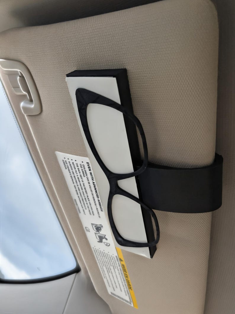 K&ouml;nnte beinhalten: Schwarze Brille mit Katzenaugen-Rahmen wird in einem schwarzen Halter an einer beigen Sonnenblende befestigt. Ein Sicherheitshinweis ist sichtbar. Der Text "EVEN WITH ADVANCED AIRBAGS" ist auf dem Etikett aufgedruckt.