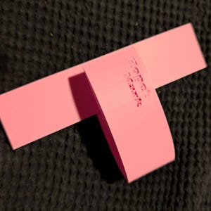 K&ouml;nnte beinhalten: Ein rosa Produktanh&auml;nger, rechteckig, mit dem gepr&auml;gten Text "HOCK MARK" auf dem gefalteten Abschnitt. Der Anh&auml;nger besteht aus einem festen, matten Material. Er liegt auf einem dunklen, strukturierten Hintergrund.