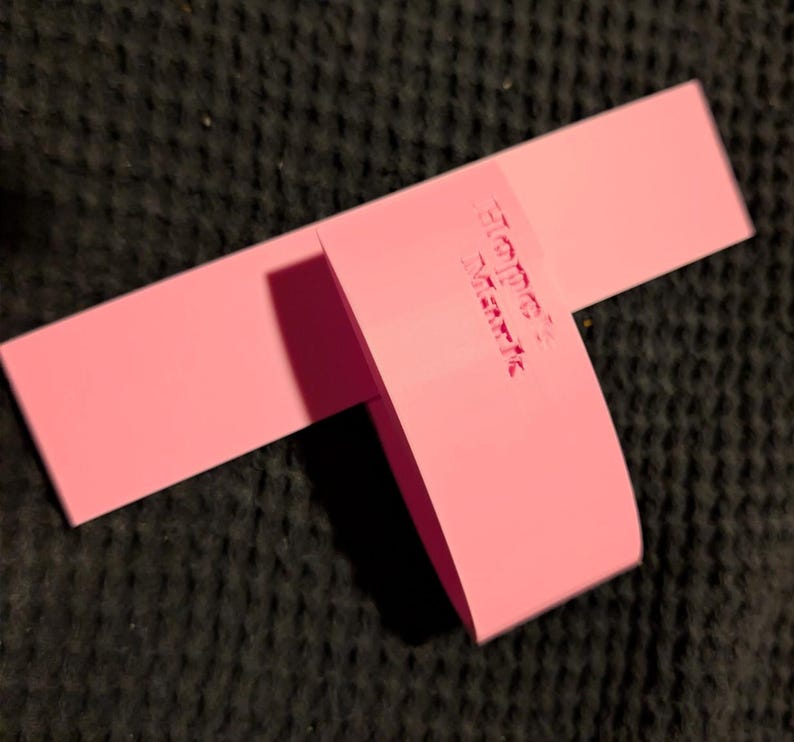K&ouml;nnte beinhalten: Ein rosa Produktanh&auml;nger, rechteckig, mit dem gepr&auml;gten Text "HOCK MARK" auf dem gefalteten Abschnitt. Der Anh&auml;nger besteht aus einem festen, matten Material. Er liegt auf einem dunklen, strukturierten Hintergrund.