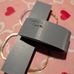 K&ouml;nnte beinhalten: Ein graues, 3D-gedrucktes Objekt mit dem Text "Hope's Market" wird gezeigt. Das Objekt ist gebogen und rechteckig und liegt auf einer rosa Oberfl&auml;che mit Herz- und Blumenmustern. Ein glitzerndes, goldfarbenes Armband ist ebenfalls zu sehen.