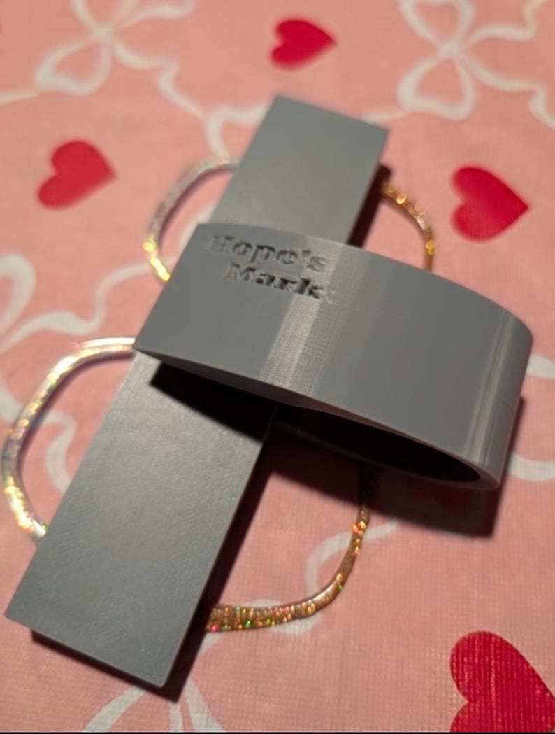 K&ouml;nnte beinhalten: Ein graues, 3D-gedrucktes Objekt mit dem Text "Hope's Market" wird gezeigt. Das Objekt ist gebogen und rechteckig und liegt auf einer rosa Oberfl&auml;che mit Herz- und Blumenmustern. Ein glitzerndes, goldfarbenes Armband ist ebenfalls zu sehen.