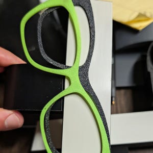 K&ouml;nnte beinhalten: Eine Brille mit einem leuchtend gr&uuml;nen Rahmen und einem strukturierten schwarzen Innenrahmen. Die Brille ist vor einem wei&szlig;en und schwarzen Hintergrund platziert, wobei eine Hand teilweise sichtbar ist, die ein schwarzes Objekt h&auml;lt.