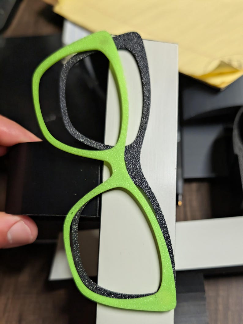 K&ouml;nnte beinhalten: Eine Brille mit einem leuchtend gr&uuml;nen Rahmen und einem strukturierten schwarzen Innenrahmen. Die Brille ist vor einem wei&szlig;en und schwarzen Hintergrund platziert, wobei eine Hand teilweise sichtbar ist, die ein schwarzes Objekt h&auml;lt.