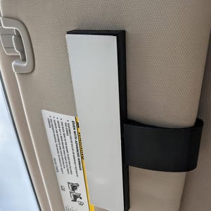 K&ouml;nnte beinhalten: Ein schwarzes und wei&szlig;es rechteckiges Objekt, das an einer Autosonnenblende befestigt ist. Das Objekt hat eine wei&szlig;e Oberfl&auml;che und einen schwarzen Rand. Die Sonnenblende ist beige, und ein Sicherheitsetikett ist sichtbar. Das Etikett enth&auml;lt den Text "EVEN WITH ADVANCED AIRBAGS".