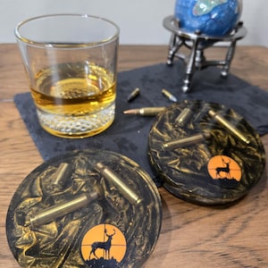 May include: Two round black and gold resin coasters with brass bullet casings and a deer silhouette target design. A rocks glass with amber liquid sits next to the coasters. A small globe on a silver stand is in the background.