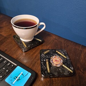 May include: A white teacup with tea sits on a black square coaster with gold accents. Another coaster with gold bullet casings and a badge is next to it. A laptop with a blue sticker is in the foreground. The background is a dark blue wall.