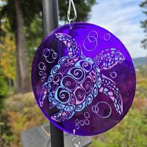 May include: A purple, circular suncatcher featuring a sea turtle design. The turtle is detailed with blue and pink swirls and bubbles. The suncatcher hangs from a silver chain with crystal accents, perfect for outdoor decor.