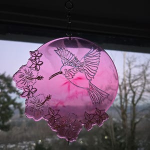 May include: A pink, circular hummingbird suncatcher with a detailed hummingbird and floral design. The suncatcher is suspended by a chain and hook. The background shows a view of trees and a cloudy sky.